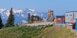 Remains of Hurricane Ridge lodge that burned down in May 2023 that shows two brick chimneys, the remains of a brick wall, and exclusion fencing and a trail around the charred remains with the Olympic Mountains looming in the background