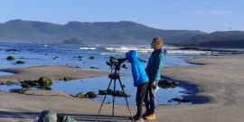 Two tour participants look through a spotting scope at birds in the Pacific Ocean with mountains in the background