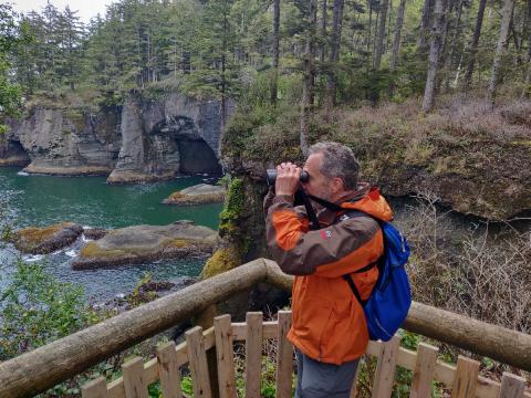 A birder looks through binoculars on one of the Cape Flattery platforms with sea caves and emerald bays in the background
