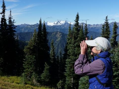 A head and shoulder shot of a birder looking through binoculars with the Olympic Mountains and Mount Olympus in the background 