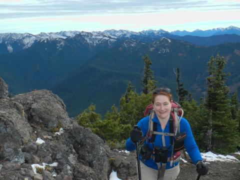 A hiker with a hydration pack, binoculars, and hiking poles smiles in the boulder field with the snowclad Olympic Mountains in the background