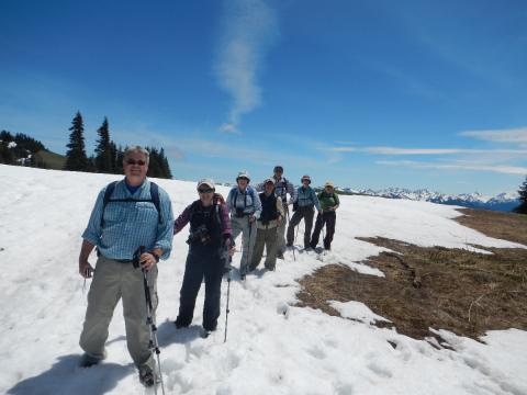 A group of six hikers take a moment to pose for the camera as they hike on snow with the snowy Olympic Mountains in the background