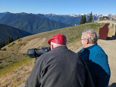 A couple looks at birds in the spotting scope from the Hurricane Ridge parking lot with the Olympic Mountains in the background