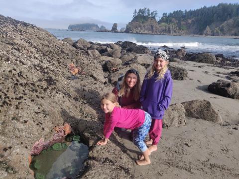 A mom and her two young girls pause for a photo next to a tidepool teeming with sea stars and anemones with seastacks and the Pacific Ocean in the background