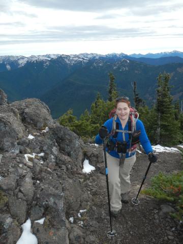 A hiker with hiking poles, a backpack, and hydration system, poses on trail dotted with snow with the Olympic Mountains in the background