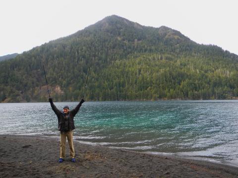 A participant stands in front of Lake Crescent with Pyramid Peak in the background with his hands and hiking poles raised in the air