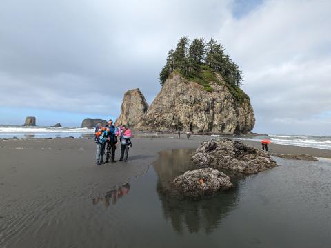 Hikers poses on a La Push beach in front of a rocky island with trees on top and rocky areas dotted with intertidal life like an orange sea star