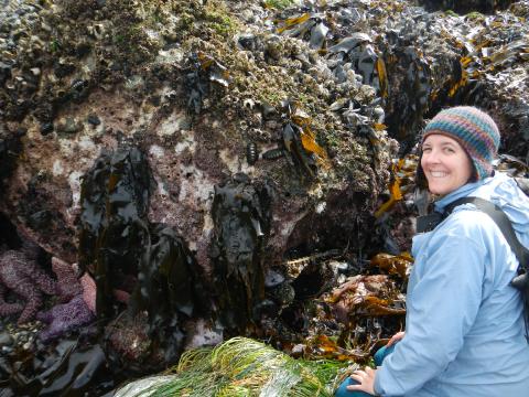 Tidepooling Guide Carolyn Poses next to a rocky outcropping covered in marine biodiversity including sea stars and seaweed during low tide