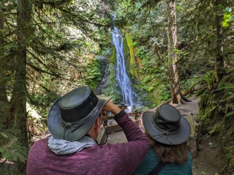 Two tour participants in brimmed summer hats take a moment to enjoy Madison Falls in the Elwha River Valley