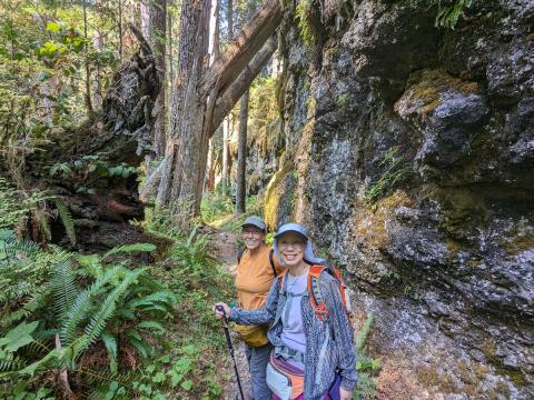 Two Women taking a moment to smile while hiking next to large rocky outcropping with teh forest, moss, and ferns in the background